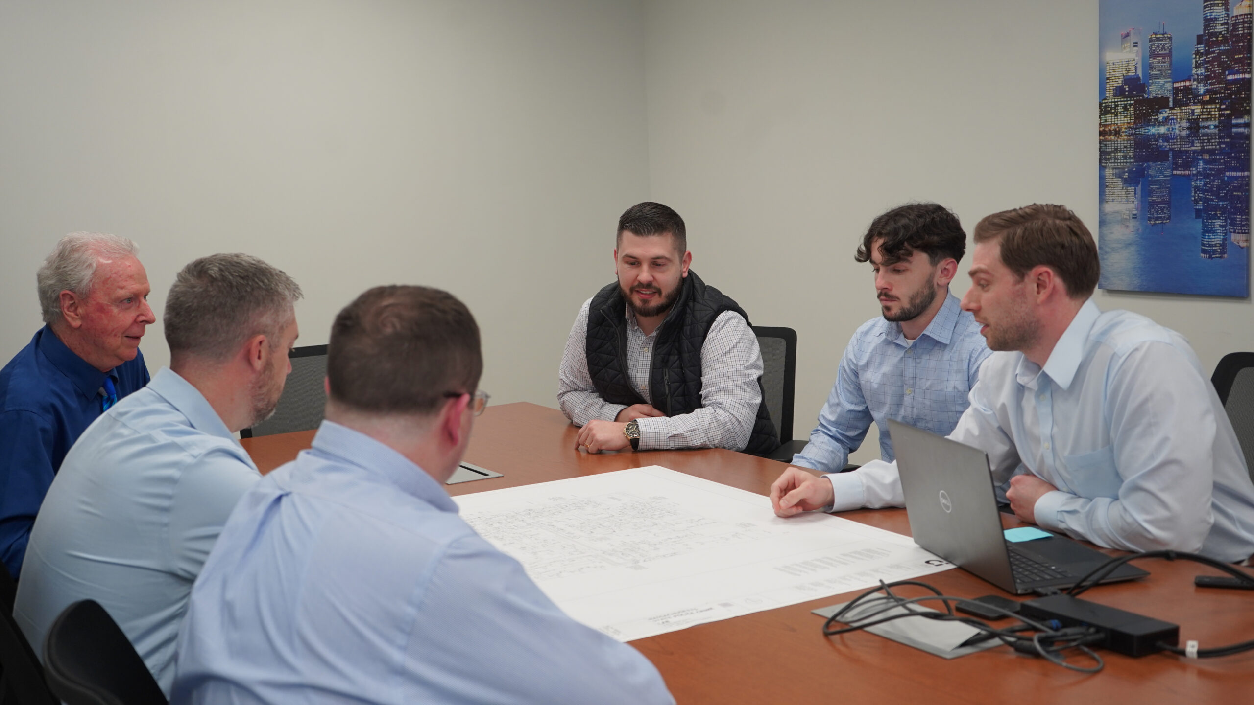 Six men sit around a conference table reviewing architectural plans, with a laptop and blueprints in front of them in a modern office setting. They appear to be discussing MEP/FP designs as part of a project for Engineered Systems Inc, ensuring every detail is meticulously planned and coordinated.