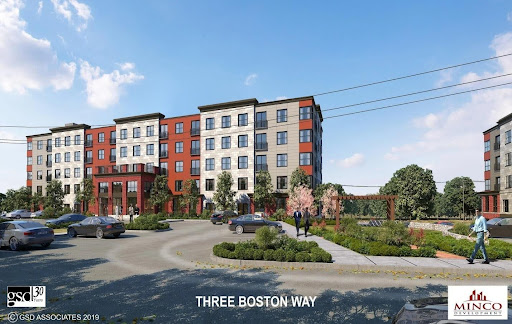 A modern five-story apartment building with a red and beige exterior, surrounded by landscaping and parked cars, labeled “THREE BOSTON WAY,” features advanced MEP/FP designs by Engineered Systems Inc for enhanced comfort and efficiency.