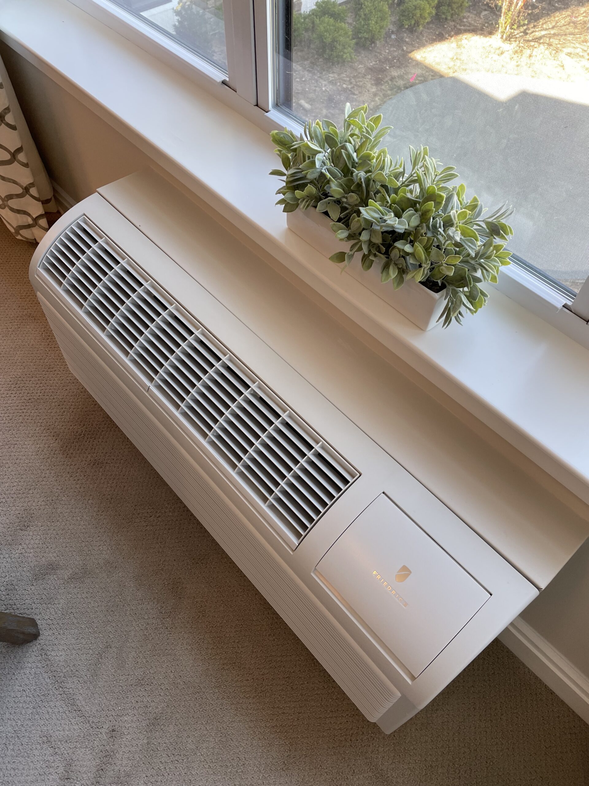 A white air conditioning unit, expertly installed below a window by Engineered Systems Inc, complements the space, with a small green potted plant placed on the windowsill above it—showcasing thoughtful MEP/FP designs for both functionality and aesthetic appeal.