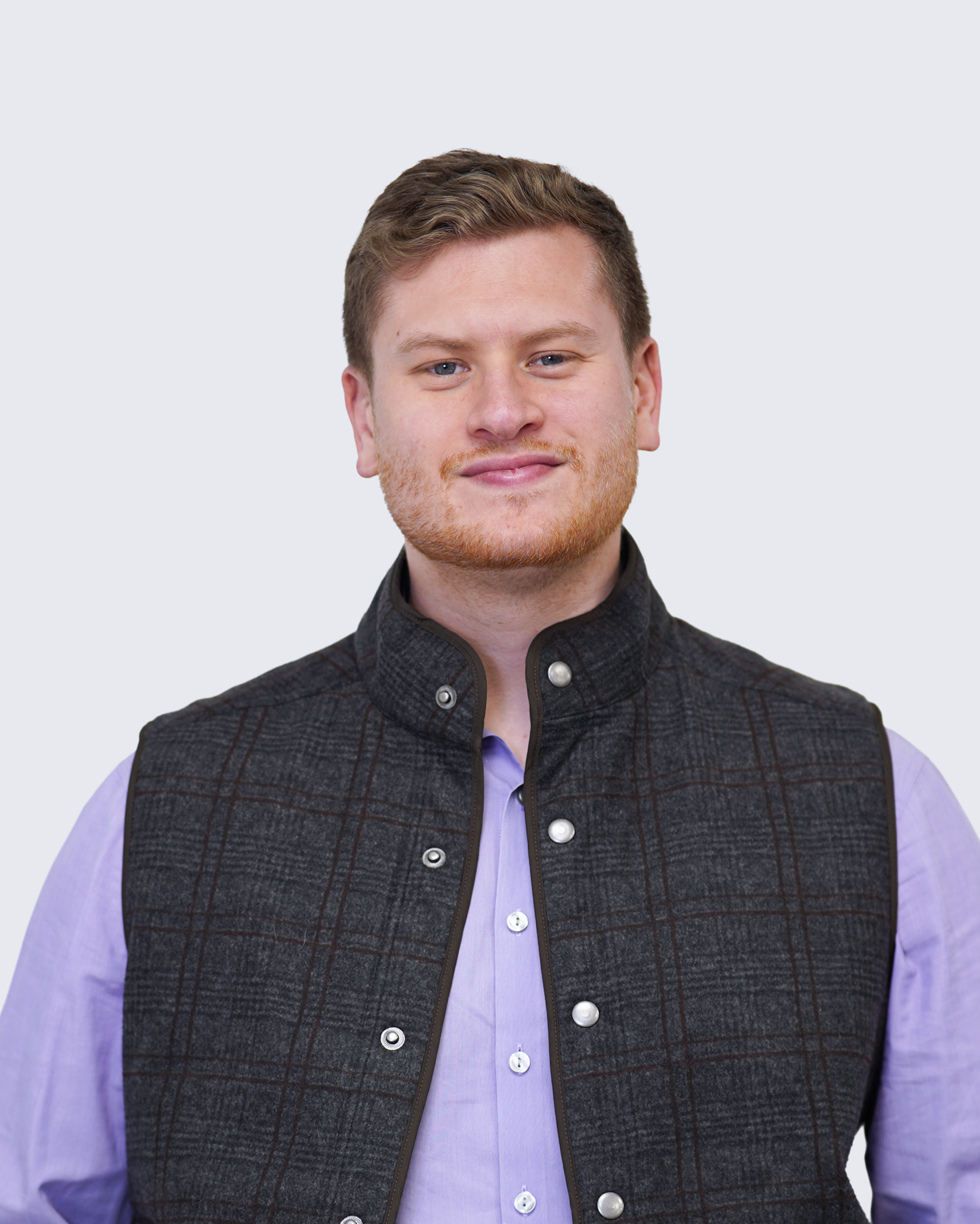 A man with short light brown hair wears a dark vest over a light purple shirt, standing against a plain light background, representing Engineered Systems Inc and their expertise in MEP/FP designs.