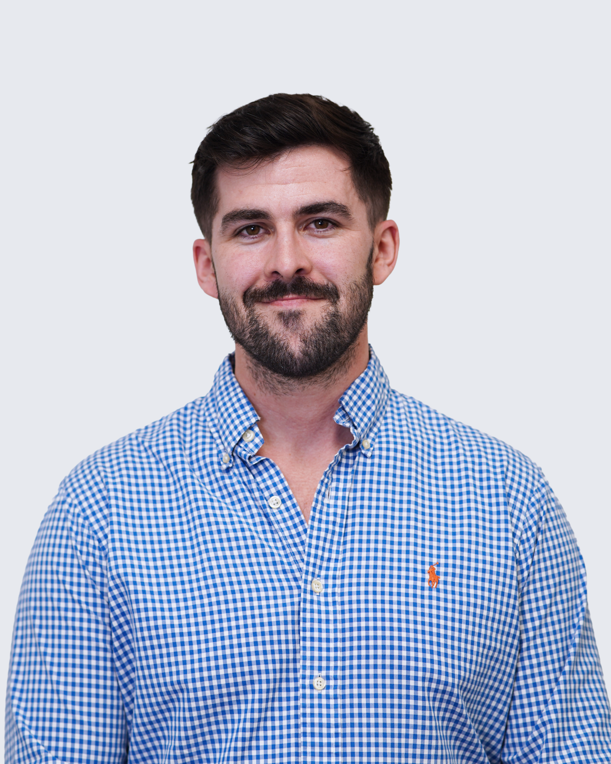 A man with short dark hair and a beard wearing a blue and white checkered shirt stands against a plain light background, embodying the professionalism associated with Engineered Systems Inc and their expertise in MEP/FP designs.