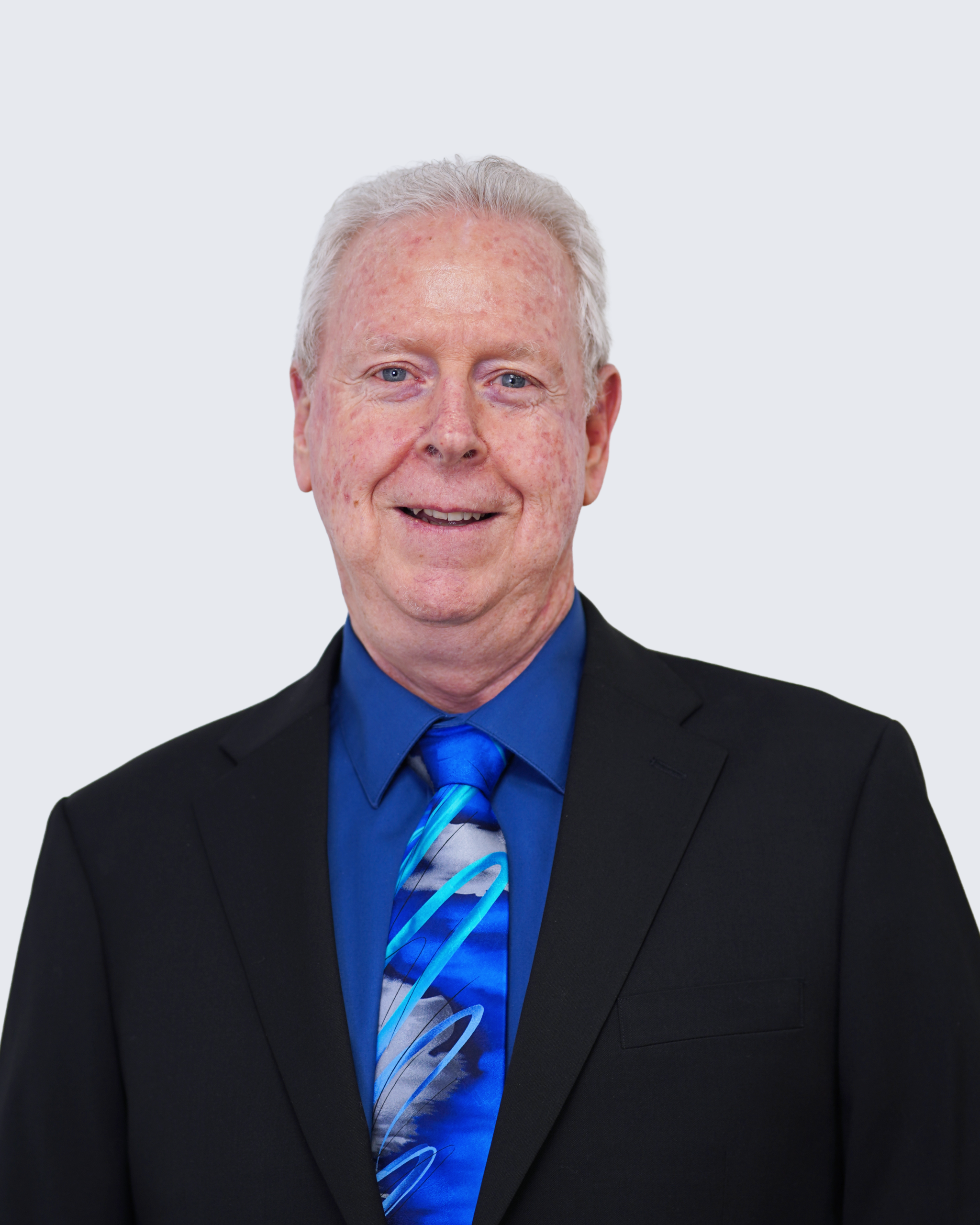 An older man with short gray hair wears a black suit, blue shirt, and blue patterned tie, standing against a plain light background—a distinguished professional representing Engineered Systems Inc and specializing in MEP/FP designs.
