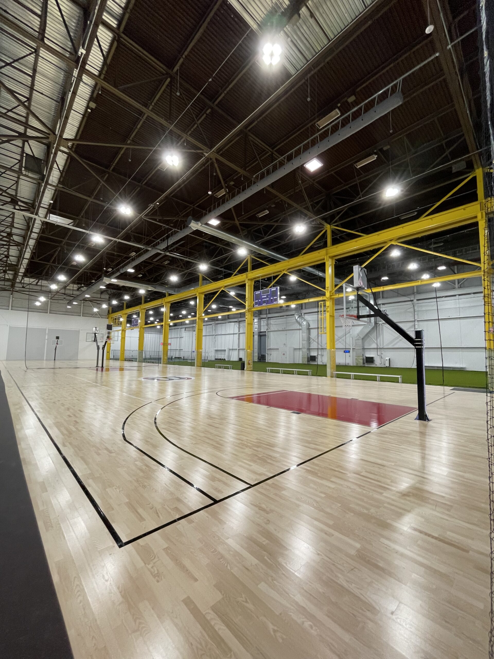 Indoor basketball court with polished wooden flooring, high ceilings, yellow steel beams, and overhead lights designed with MEP/FP designs from Engineered Systems Inc; a visible soccer field can be seen in the background.