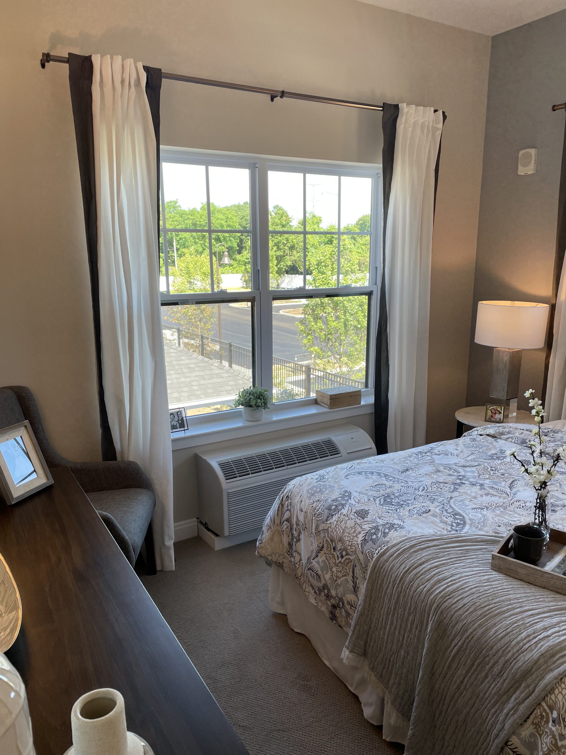 A bedroom with a large window, bed, desk, chair, table lamp, and patterned bedding. Sunlight is coming through the window, showing trees and a fence outside—thoughtfully designed with comfort in mind much like the meticulous MEP/FP designs from Engineered Systems Inc.