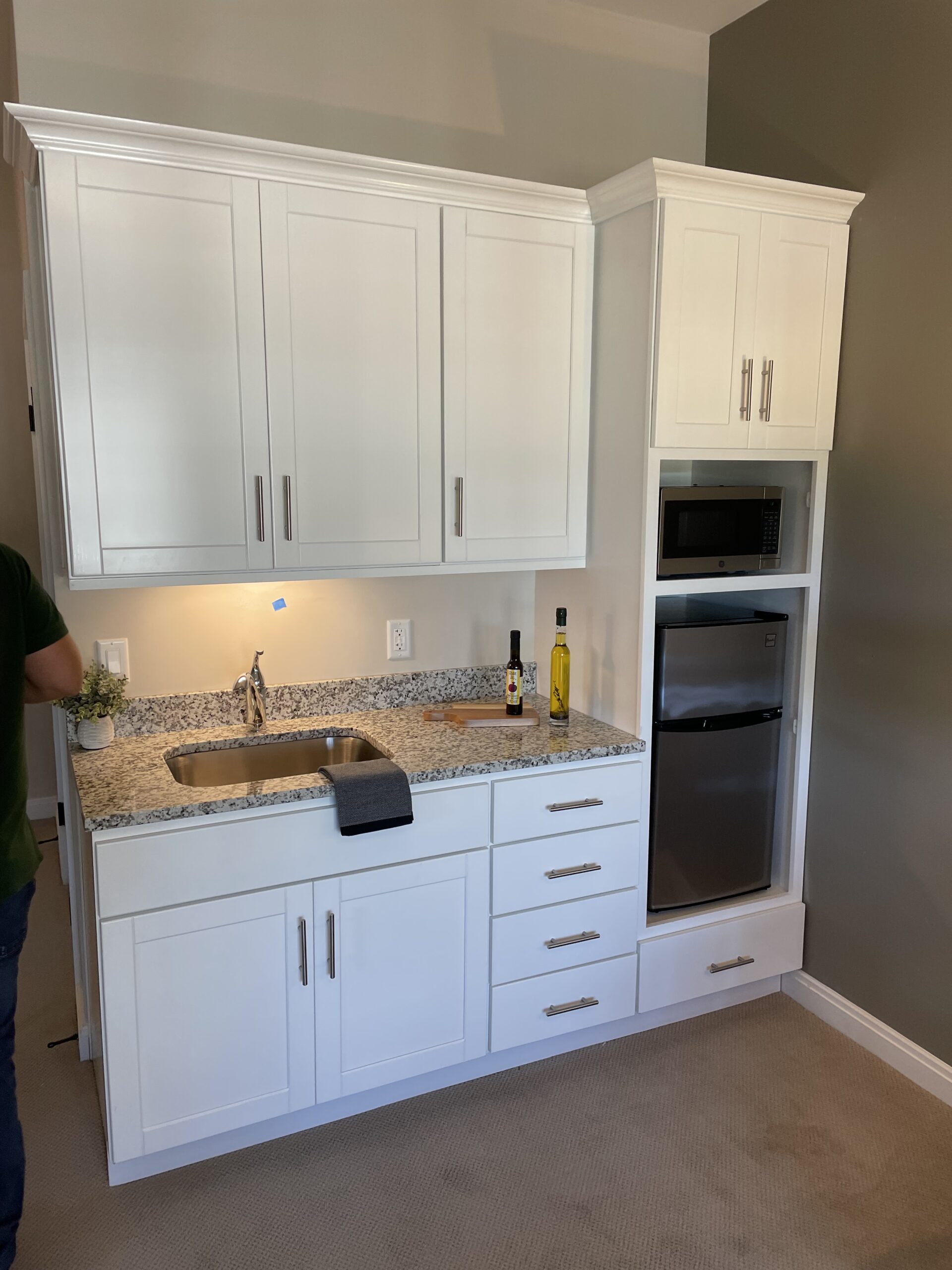 White kitchenette with granite countertop, sink, upper and lower cabinets, microwave, mini fridge, and small decor—demonstrating thoughtful MEP/FP designs from Engineered Systems Inc. Person partially visible on the left.