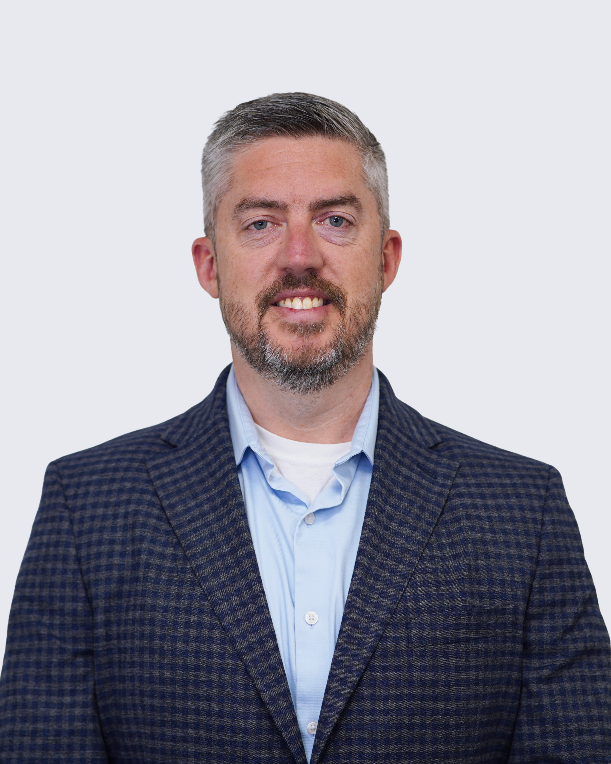Man with short gray hair and beard wearing a blue plaid blazer over a light blue shirt, standing against a plain light background, facing the camera and smiling slightly—representing Engineered Systems Inc and their expertise in MEP/FP designs.