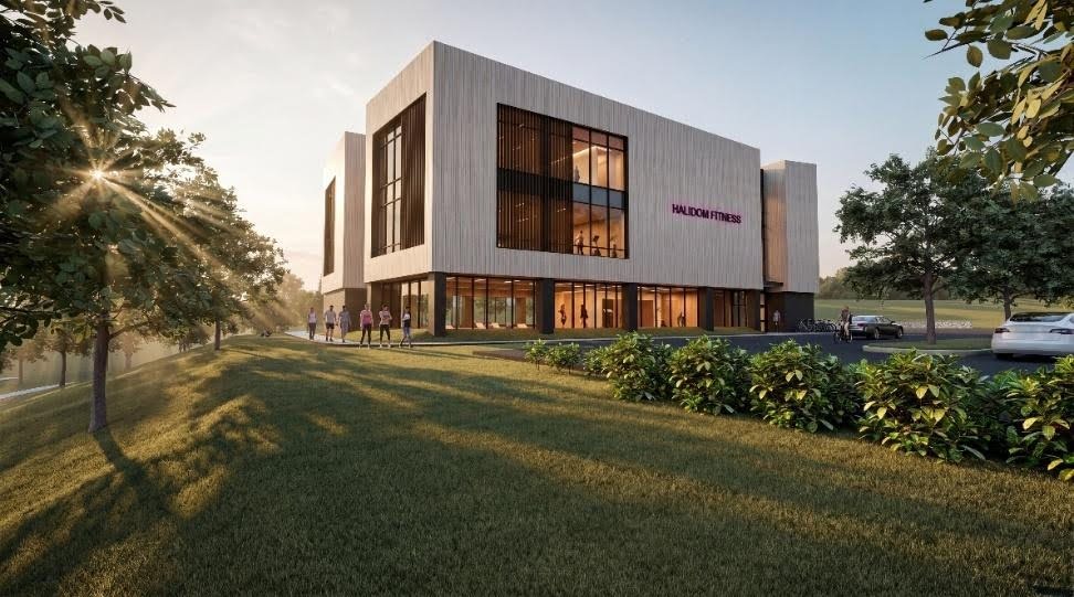 A modern, multi-story fitness center with large windows, surrounded by trees and greenery, showcases state-of-the-art MEP/FP designs by Engineered Systems Inc, as people walk toward the entrance and cars are parked nearby.