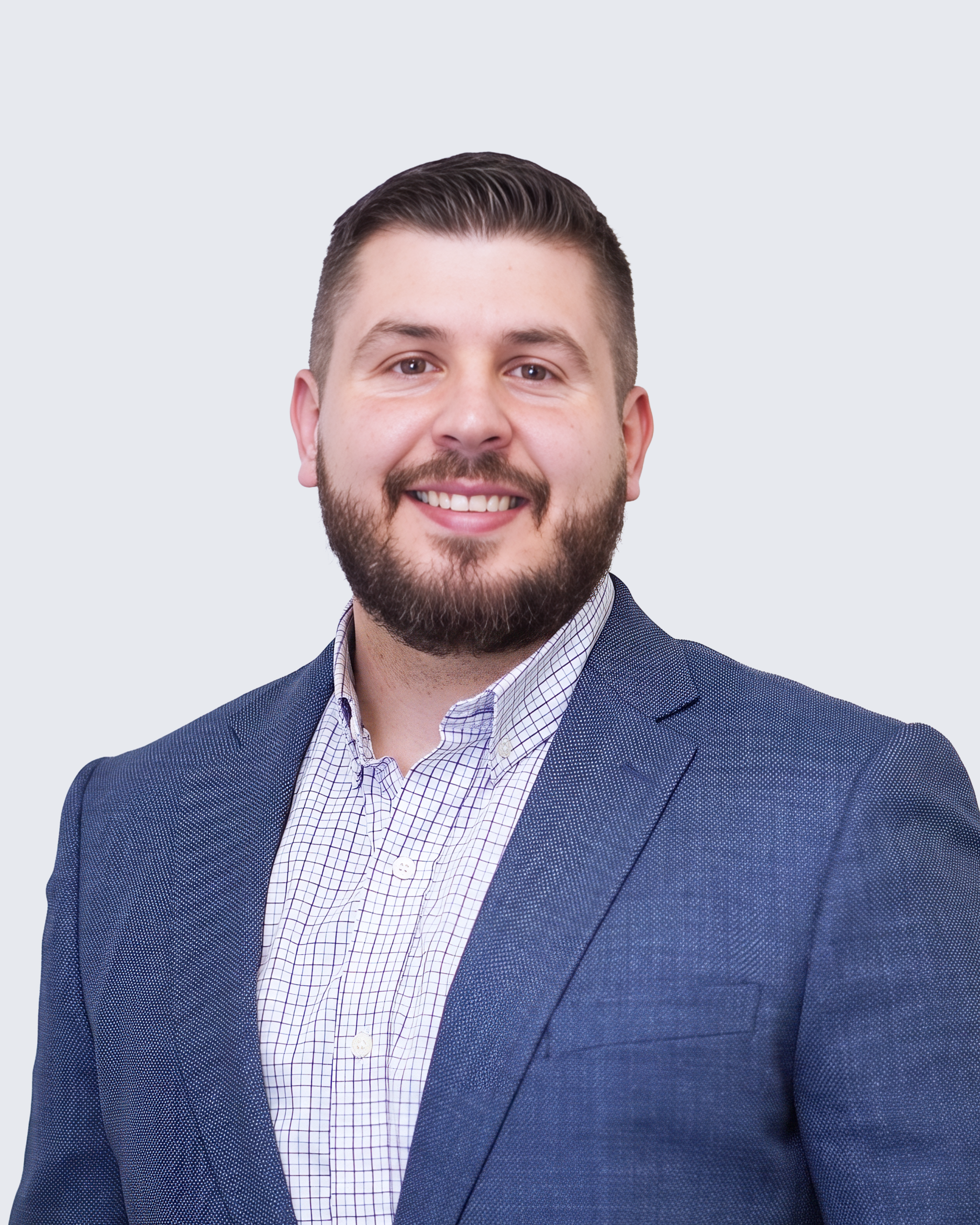 A smiling man with a beard, wearing a blue blazer and a button-down grid-patterned shirt, poses against a light grey background—reflecting the professionalism and approachability at Engineered Systems Inc, specialists in MEP/FP designs.