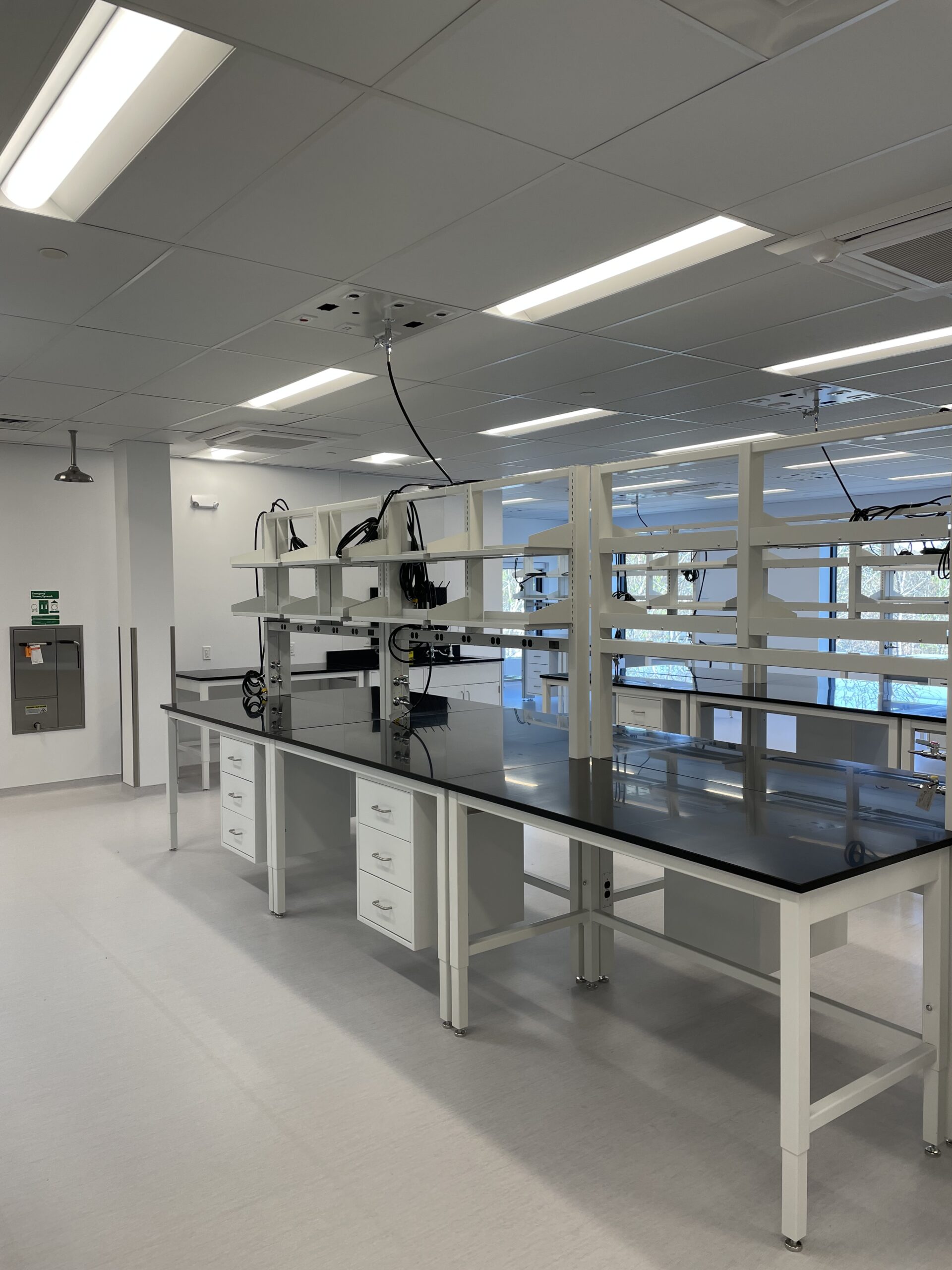 A clean, empty laboratory with black workbenches, white shelves, storage drawers, and ceiling-mounted equipment—expertly designed by Engineered Systems Inc using innovative MEP/FP designs. Bright lighting and large windows provide natural light.