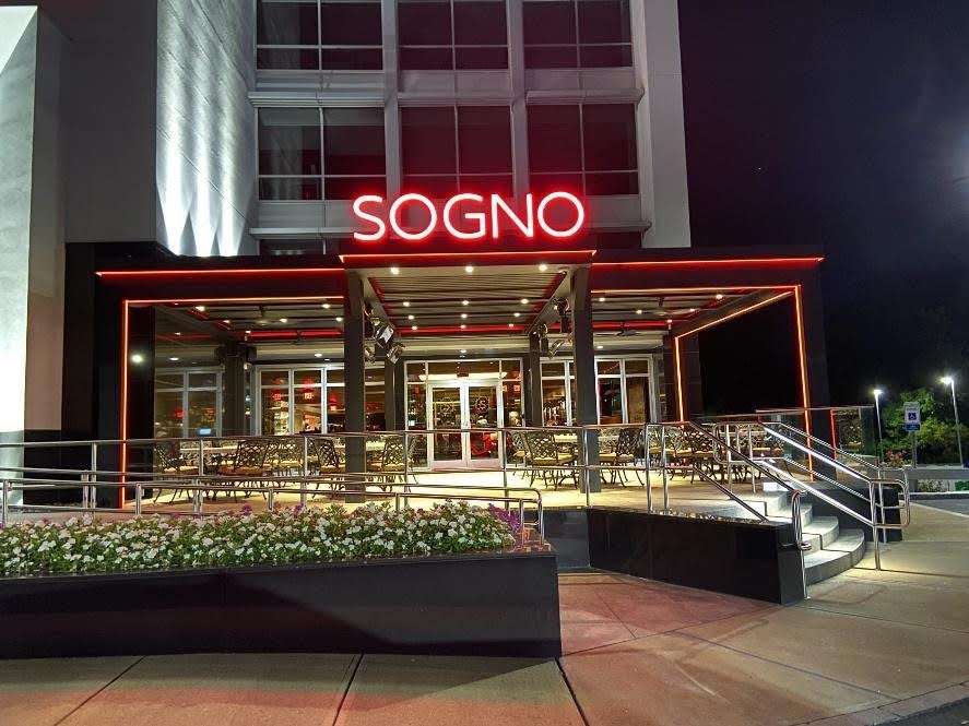 Exterior of a restaurant named SOGNO at night, featuring red neon lighting, glass doors, outdoor seating, and a ramp for accessibility—showcasing thoughtfully integrated MEP/FP designs by Engineered Systems Inc.